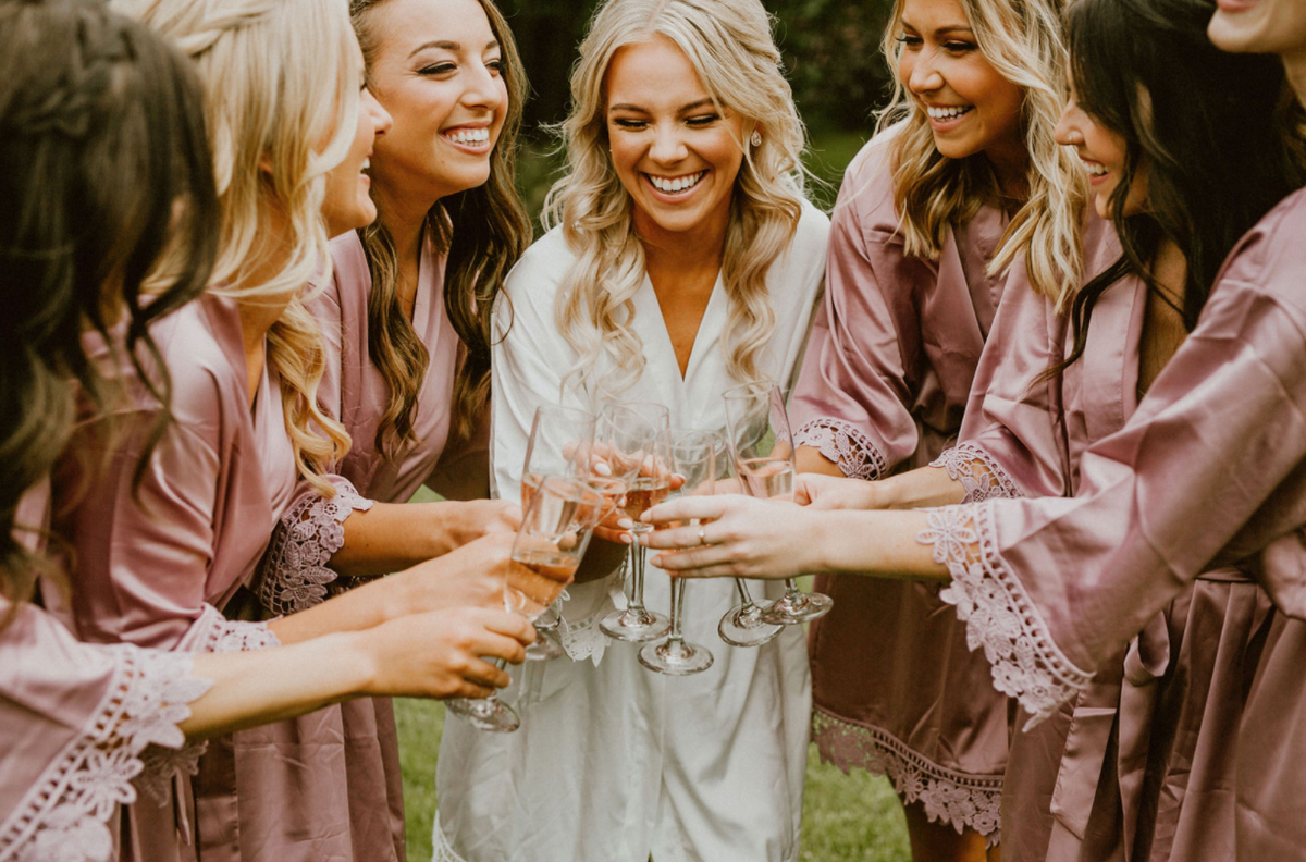 Bridal party wearing Aurora satin robes in white and mauve- cheers with champagne glasses. 
